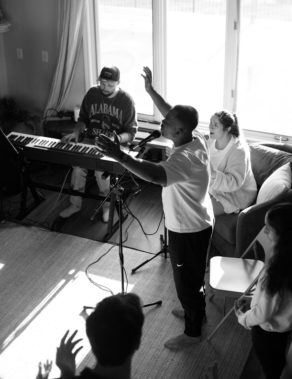 A worship leader lifts his hand as a keyboard player leads music in a sunlit living room